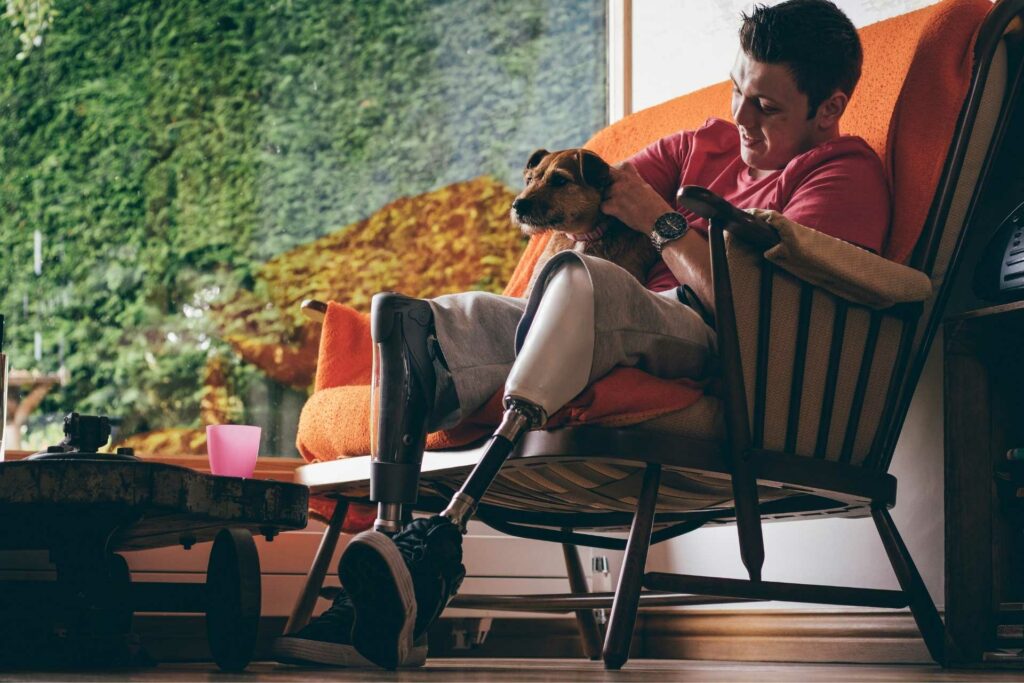 A young man with quadriplegia sitting on a wooden chair patting a small scruffy dog on his lap.